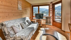 A cozy room with a wooden wall, a patterned sofa, a television, and a view of mountains through large windows. - Les Gets - 2BR Apartment for 6 with Balcony and WIFI (Les Gets)