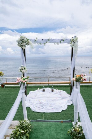 An outdoor wedding arch decorated with flowers and draped in white fabric, overlooking a body of water.