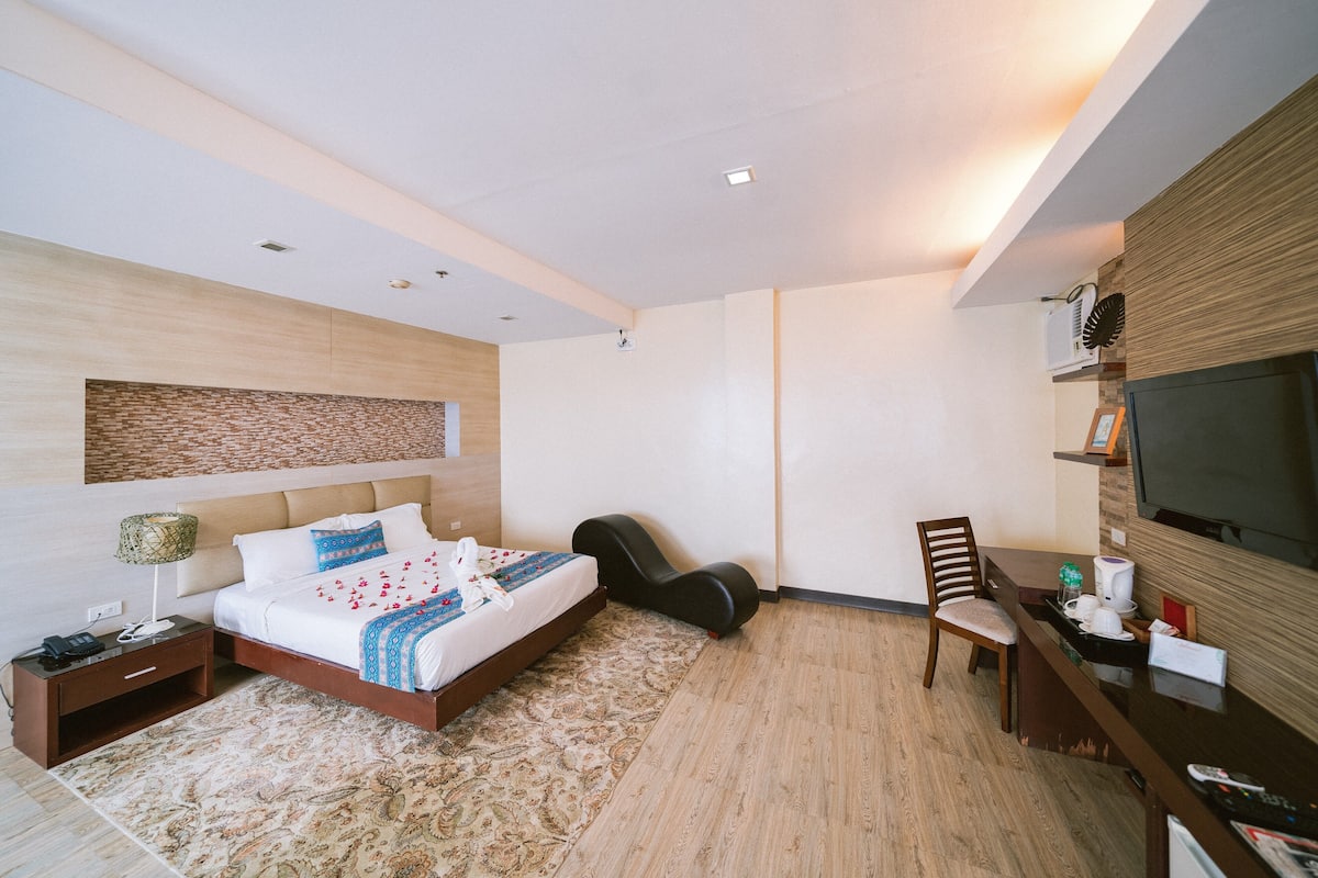 A modern hotel room with a bed, bedside table, chair, desk, and TV.