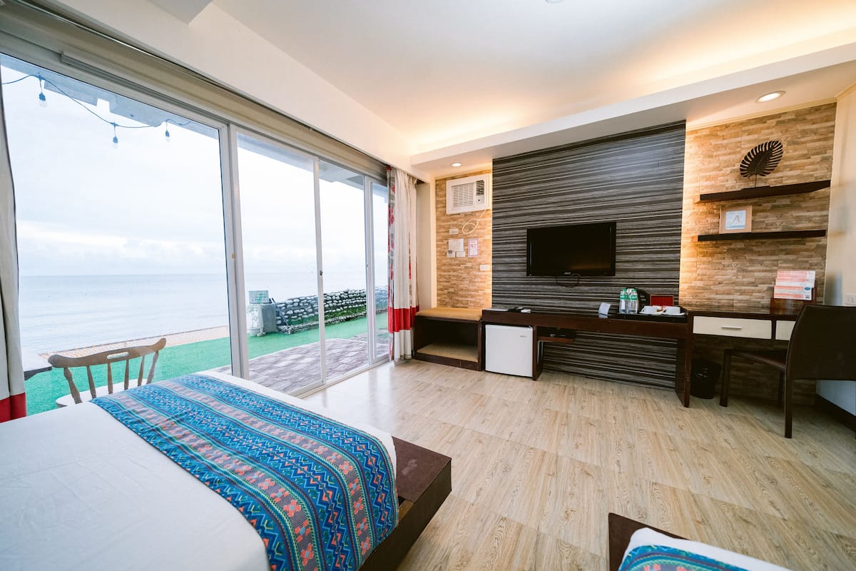 A modern hotel room with a large bed, a desk, a flat-screen TV, and a balcony with a view of the sea.