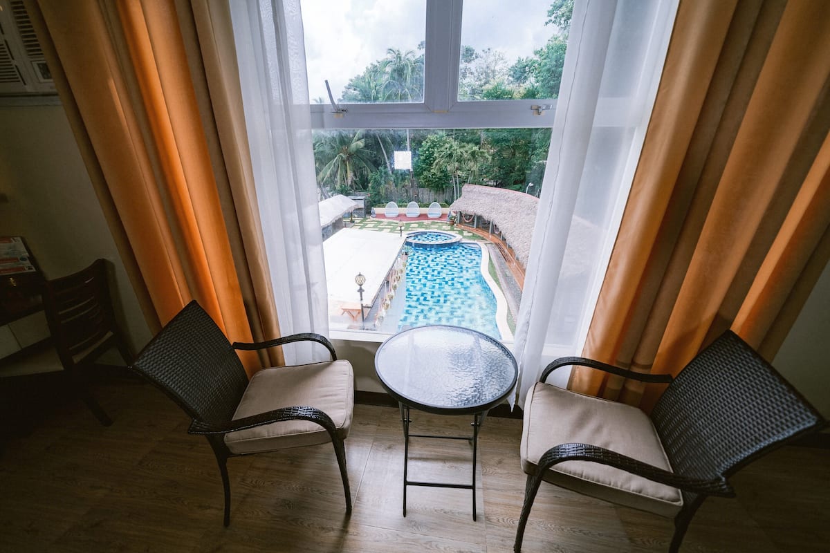 A room with two wicker chairs and a glass table, overlooking a swimming pool with a thatched roof area.