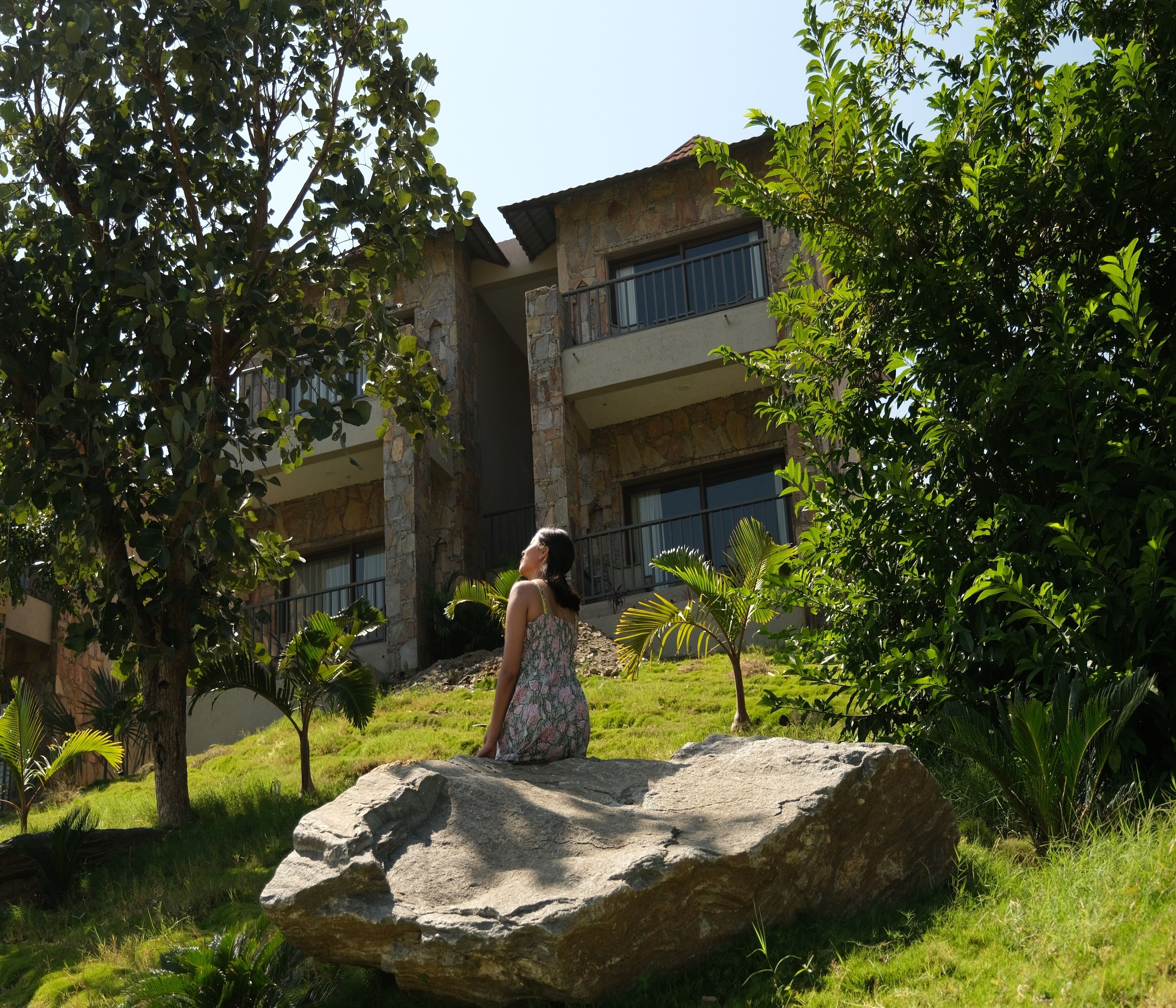 A woman sitting on a rock in a lush garden with a building in the background.