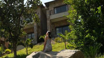 A woman sitting on a rock in a lush garden with a building in the background.
