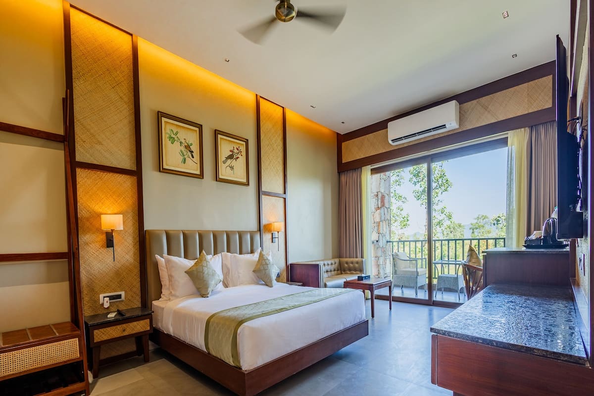 A hotel room with a large bed, a desk, a balcony with a table and chairs, and framed artwork on the wall.