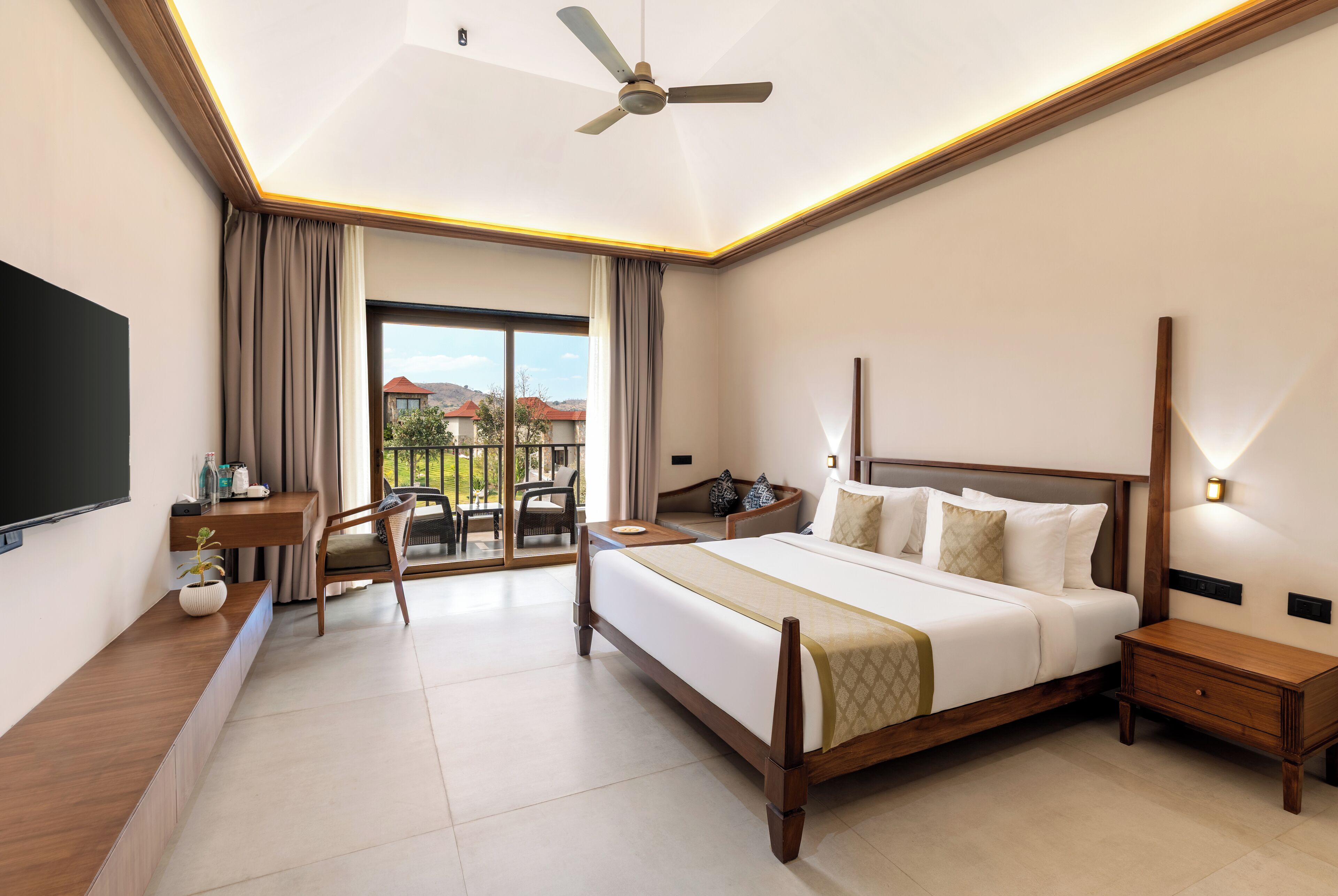 A hotel room with a bed, a desk, a chair, a TV, a ceiling fan, and a view of the outdoors.