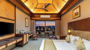 A hotel room with a bed, a desk, a TV, a ceiling fan, and a view of houses in the distance. - Sajjan Bagh Resort Kumbhalgarh (Kumbhalgarh)