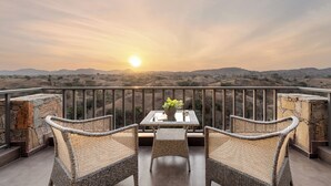A balcony with wicker furniture and a table, overlooking a mountainous landscape during sunset. - Sajjan Bagh Resort Kumbhalgarh (Kumbhalgarh)