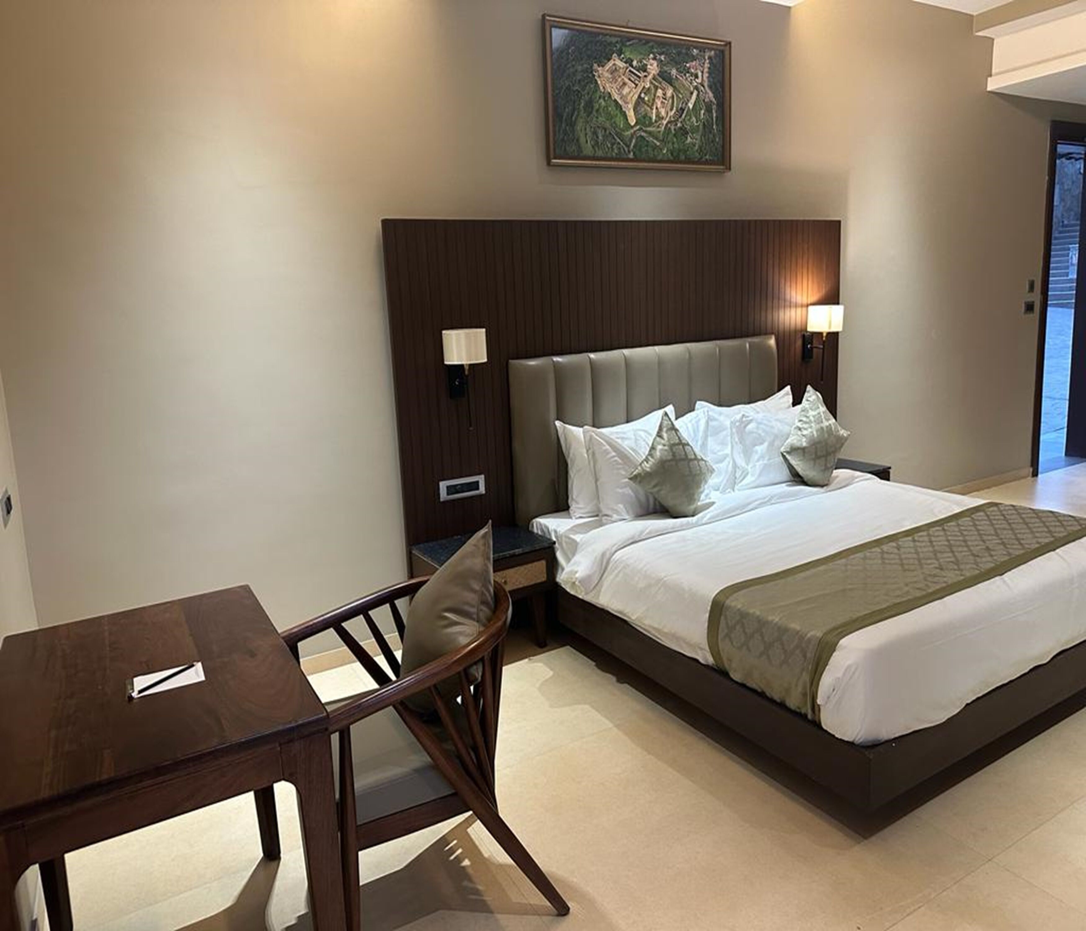 A hotel room with a large bed, a desk, and a chair. There is a painting on the wall above the bed.