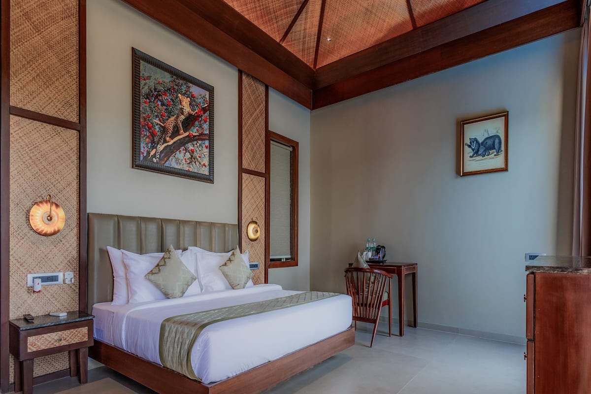 A bedroom with a large bed, a wooden headboard, a nightstand, a desk, and a chair. There are wall-mounted lights and a painting above the bed.