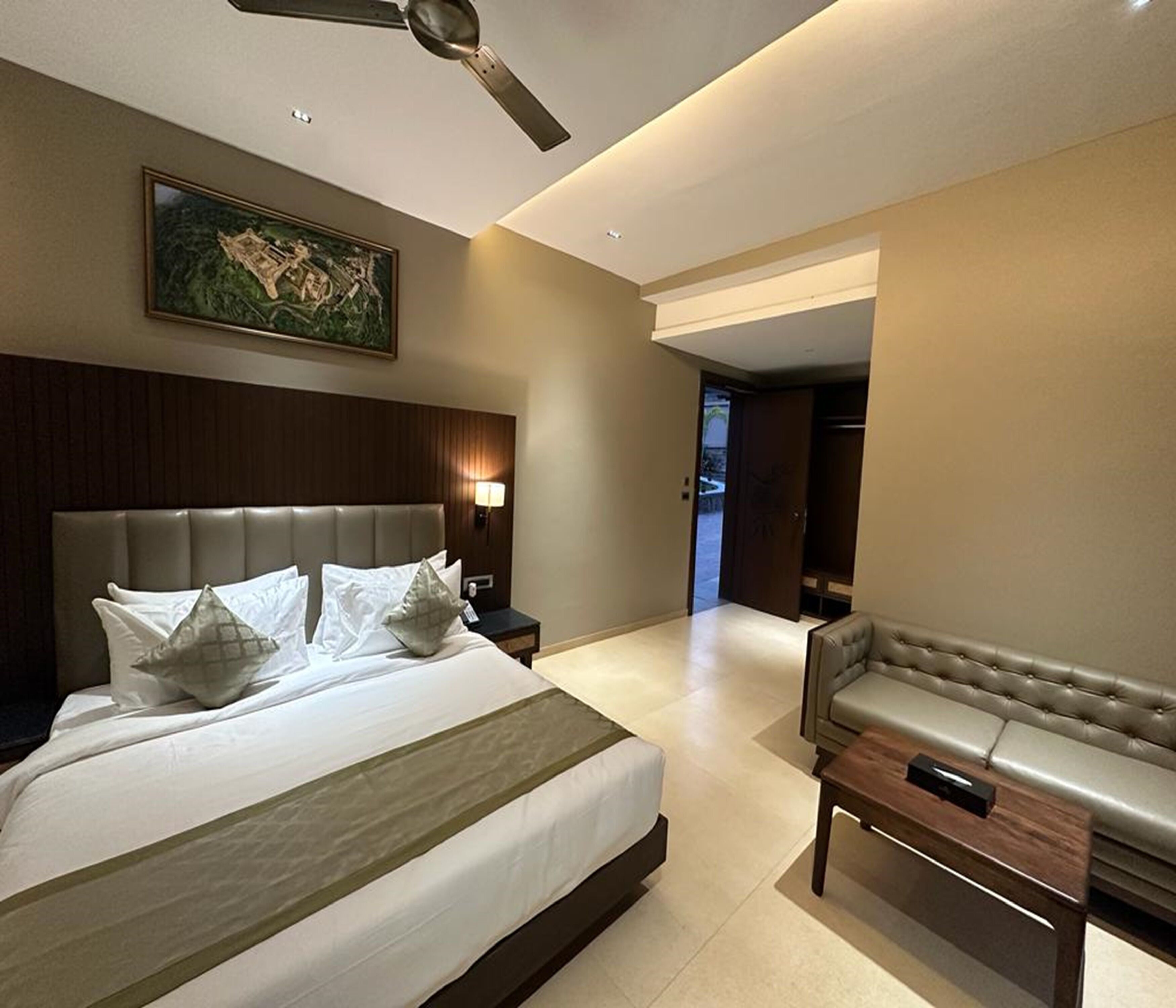 A modern hotel room with a large bed, a seating area with a sofa, a ceiling fan, and a painting on the wall.