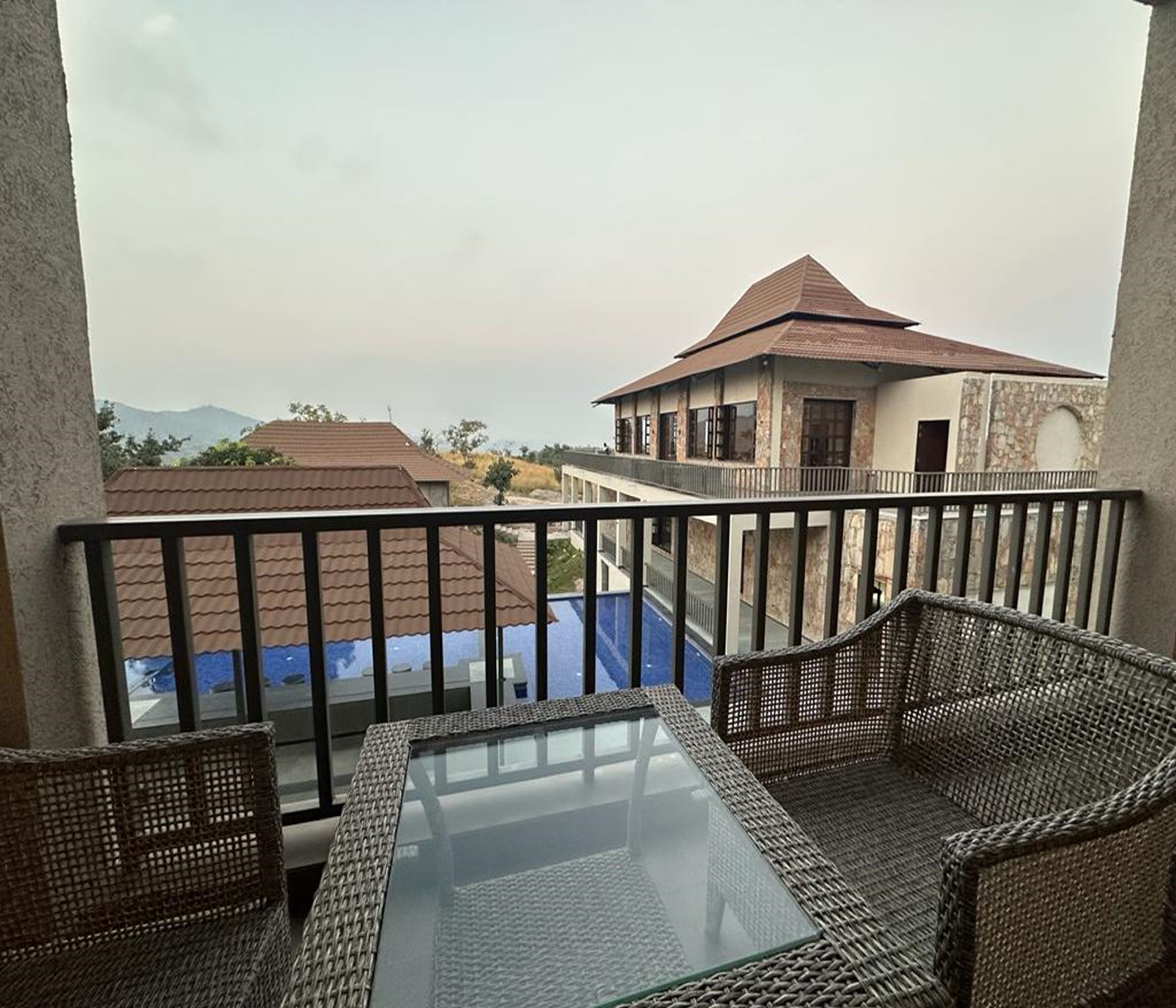 Ein Balkon mit Korbmöbeln und einem Glastisch, mit Blick auf ein Gebäude mit rotem Dach und eine ferne Berglandschaft.