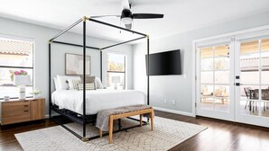 A modern bedroom with a four-poster bed, a ceiling fan, a TV mounted on the wall, and a view of the outdoor patio through a sliding glass door.