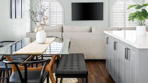 A modern kitchen with a dining area, featuring a glass table, black chairs, a grey sofa, and a large TV mounted on the wall.