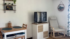 A small, well-lit room with a wooden dining table, chairs, a TV on a white cabinet, and a white armchair. - Studio à Gréoux les Bains - Les Fontaines (Gréoux-les-Bains)