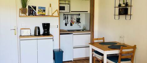 Een kleine keuken met witte kastjes, een houten eettafel met stoelen en een blauwe prullenbak.