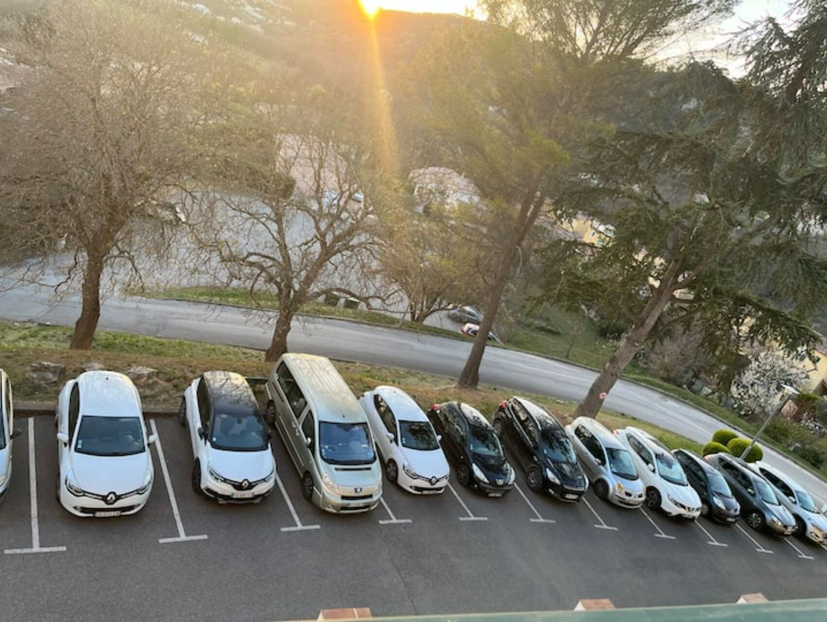A parking lot with multiple cars parked in rows, surrounded by trees and a road in the background.