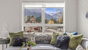 A living room with a large window offering a view of mountains and a building. There is a sofa with various cushions and a vase with flowers.