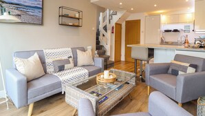 Un salon moderne avec un canapé gris, une table basse et deux fauteuils. Un escalier mène à un niveau supérieur.