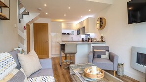 Un salon moderne avec un canapé, des fauteuils, une table basse et une cheminée. On aperçoit une cuisine à l’arrière-plan, avec un coin repas.