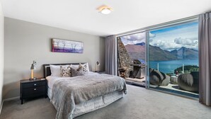 Een slaapkamer met een groot bed, een nachtkastje, een lamp, een schilderij en uitzicht op bergen en een meer door een glazen deur.