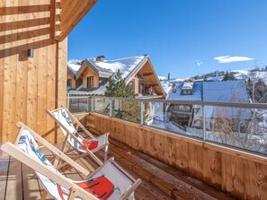 A wooden deck with two lounge chairs, a glass balustrade, and a view of snow-covered houses.