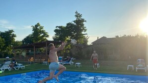 A swimming pool with people playing, a grassy area with lounge chairs, and a clear sky.