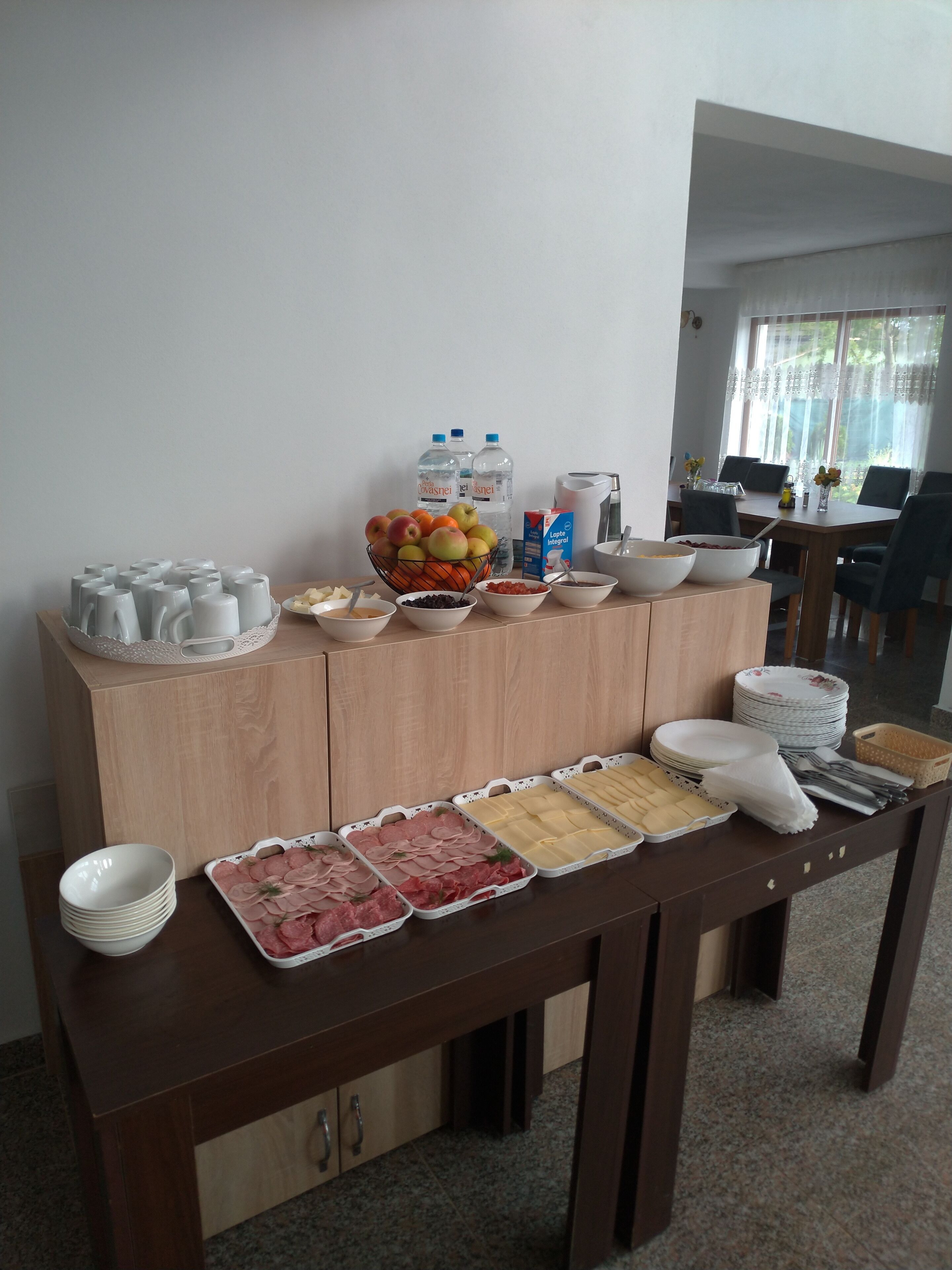 Ein reichhaltiges Buffet mit verschiedenen Speisen, Tellern und Schüsseln auf einem Holztisch, mit einem Fenster und einem Essbereich im Hintergrund.