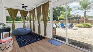 Een kamer met een plafondventilator, een kruipzak, een tafeltje met een lamp, en uitzicht op een terras met stoelen en planten.