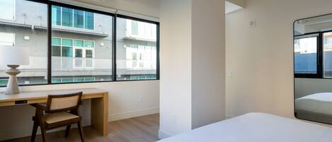 A modern hotel room with a large bed, a desk with a chair, and a large window offering a view of the building's exterior.