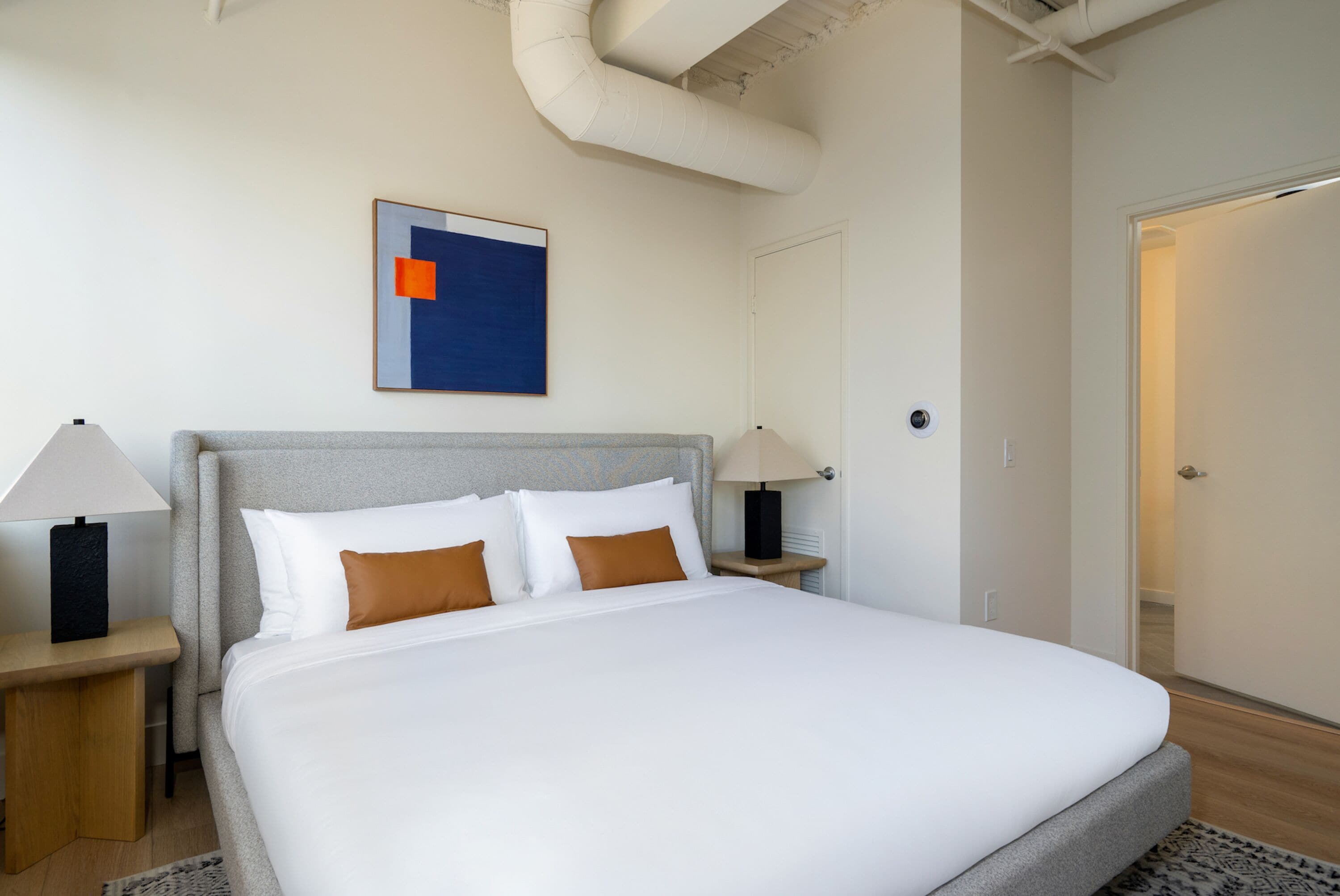 Una cama bien hecha con ropa de cama blanca y marrón, dos mesitas de noche con lámparas, una obra de arte abstracta enmarcada en la pared y una puerta visible a la derecha.
