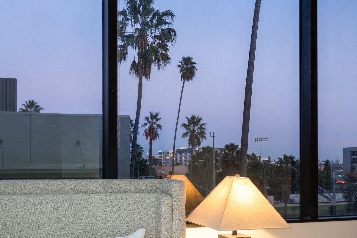 A hotel room with a bed, a nightstand with a lamp, and a large window with a view of palm trees and buildings.