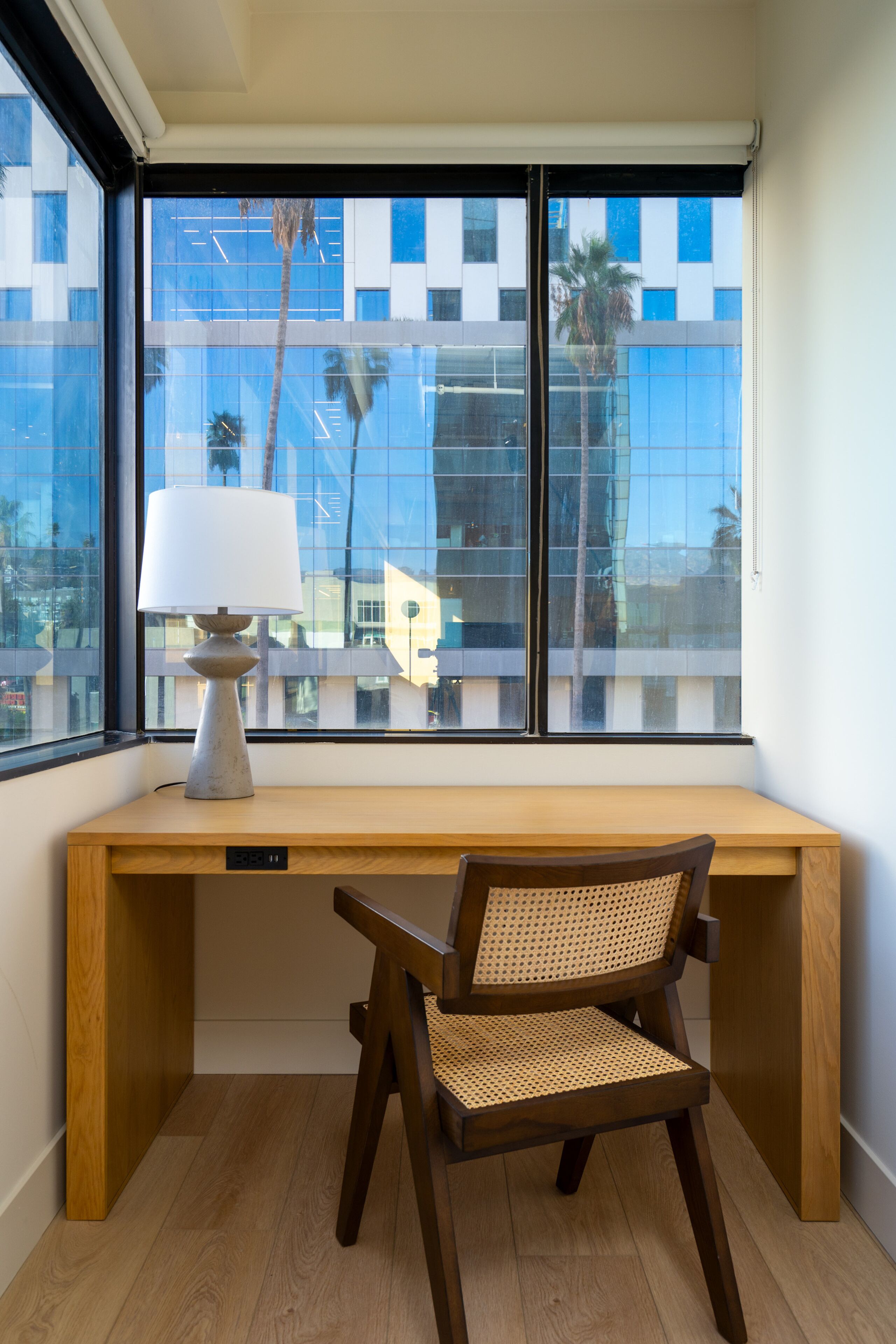 Un bureau d’angle avec une lampe, une chaise et une vue sur un immeuble moderne par la fenêtre.