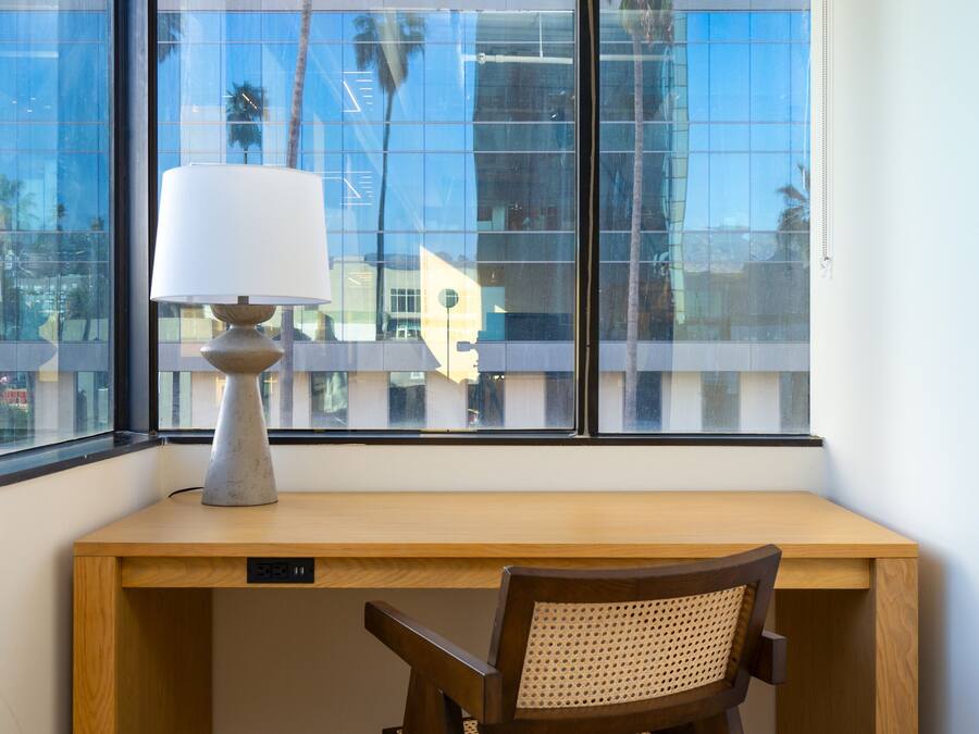 Un bureau d’angle avec une lampe, une chaise et une vue sur un immeuble moderne par la fenêtre.