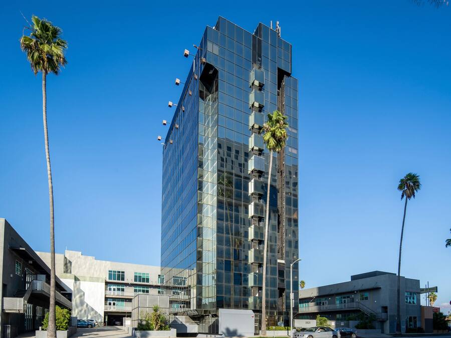 A modern high-rise building with a glass facade, surrounded by palm trees and other buildings.
