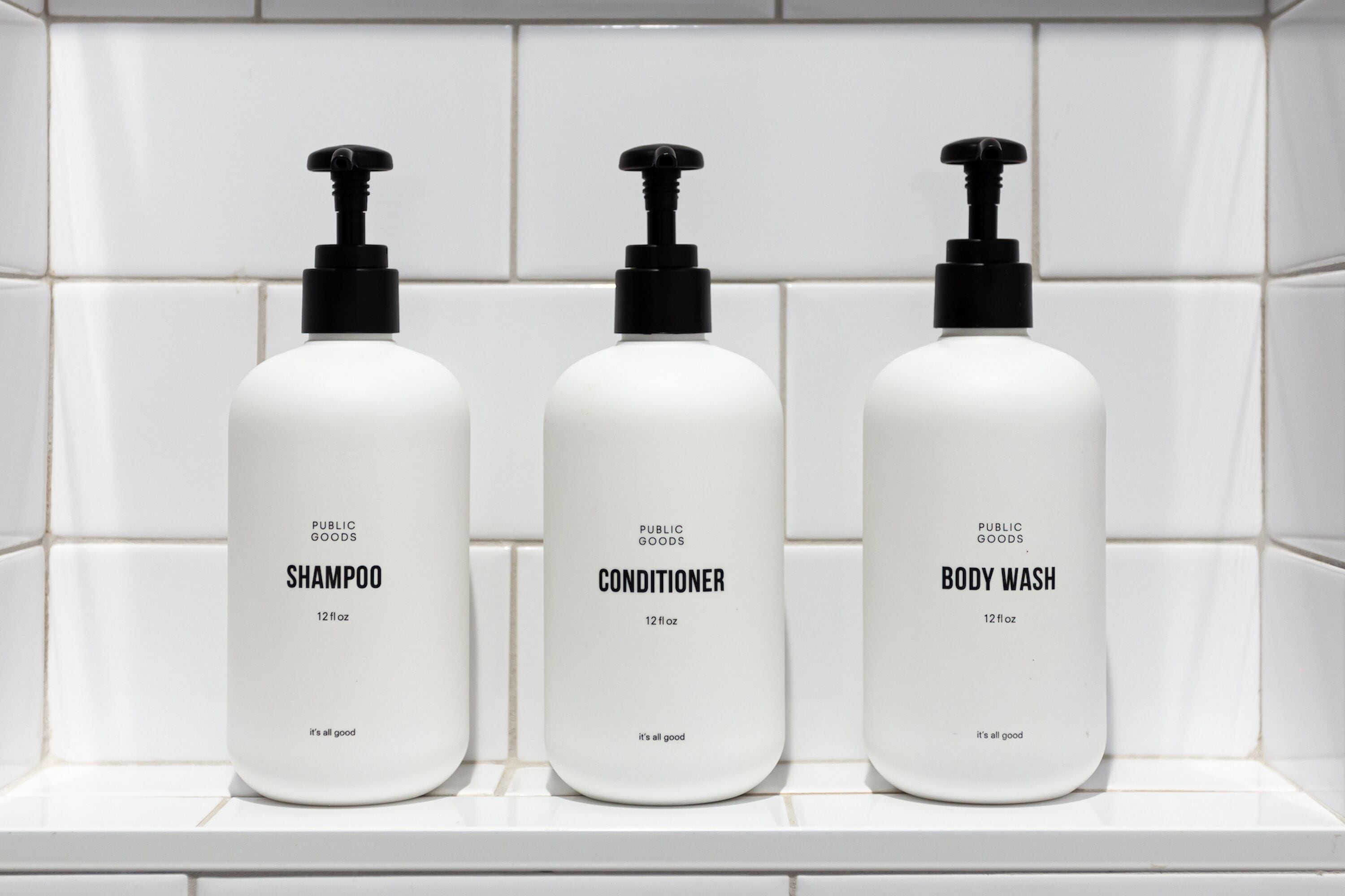 Three bottles of public goods shampoo, conditioner, and body wash on a white tiled shelf.