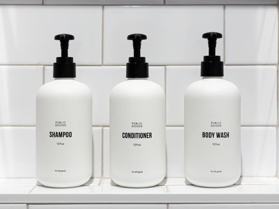 Three bottles of public goods shampoo, conditioner, and body wash on a white tiled shelf.
