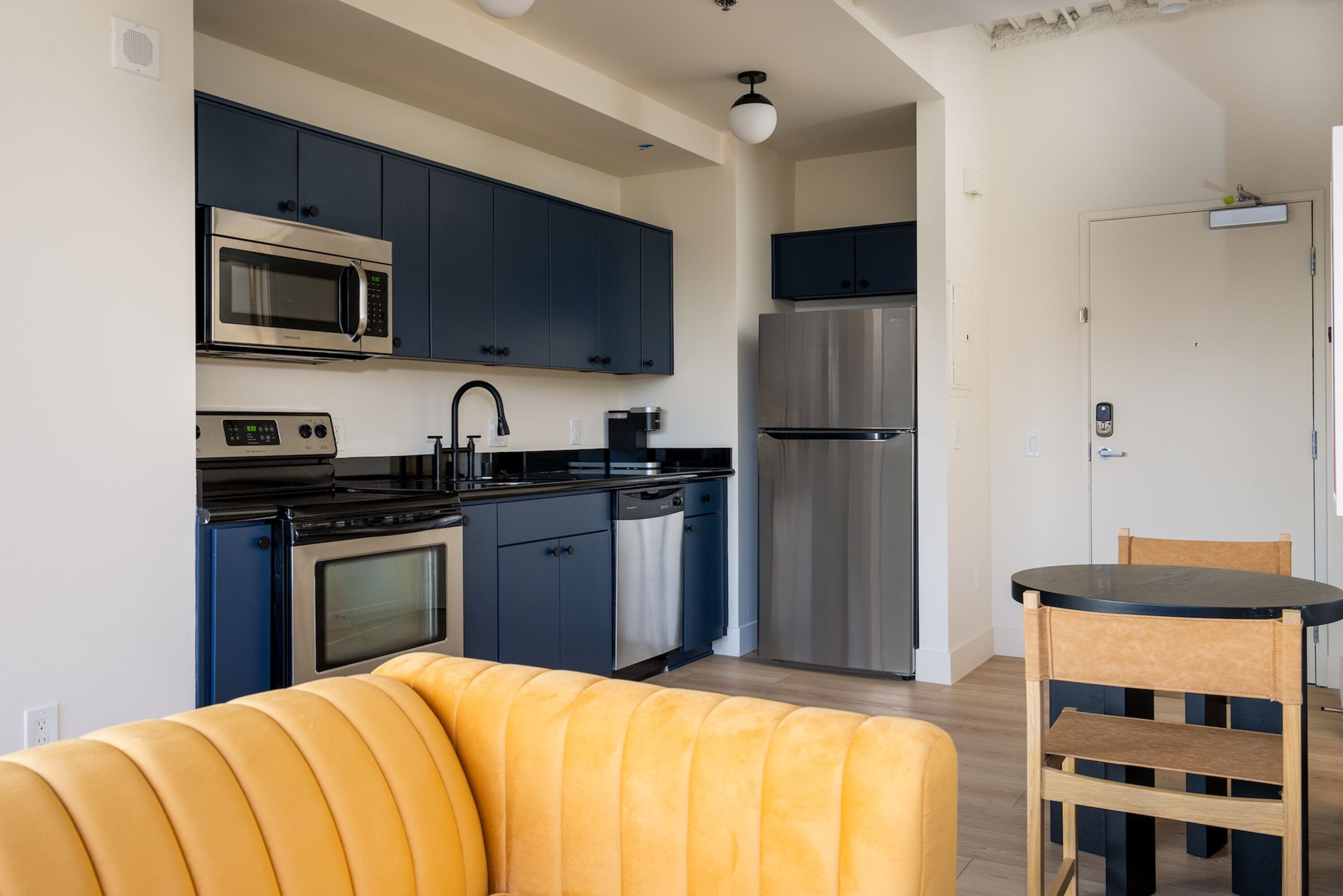 A modern kitchen with dark blue cabinets, stainless steel appliances, and a yellow sofa in the foreground.