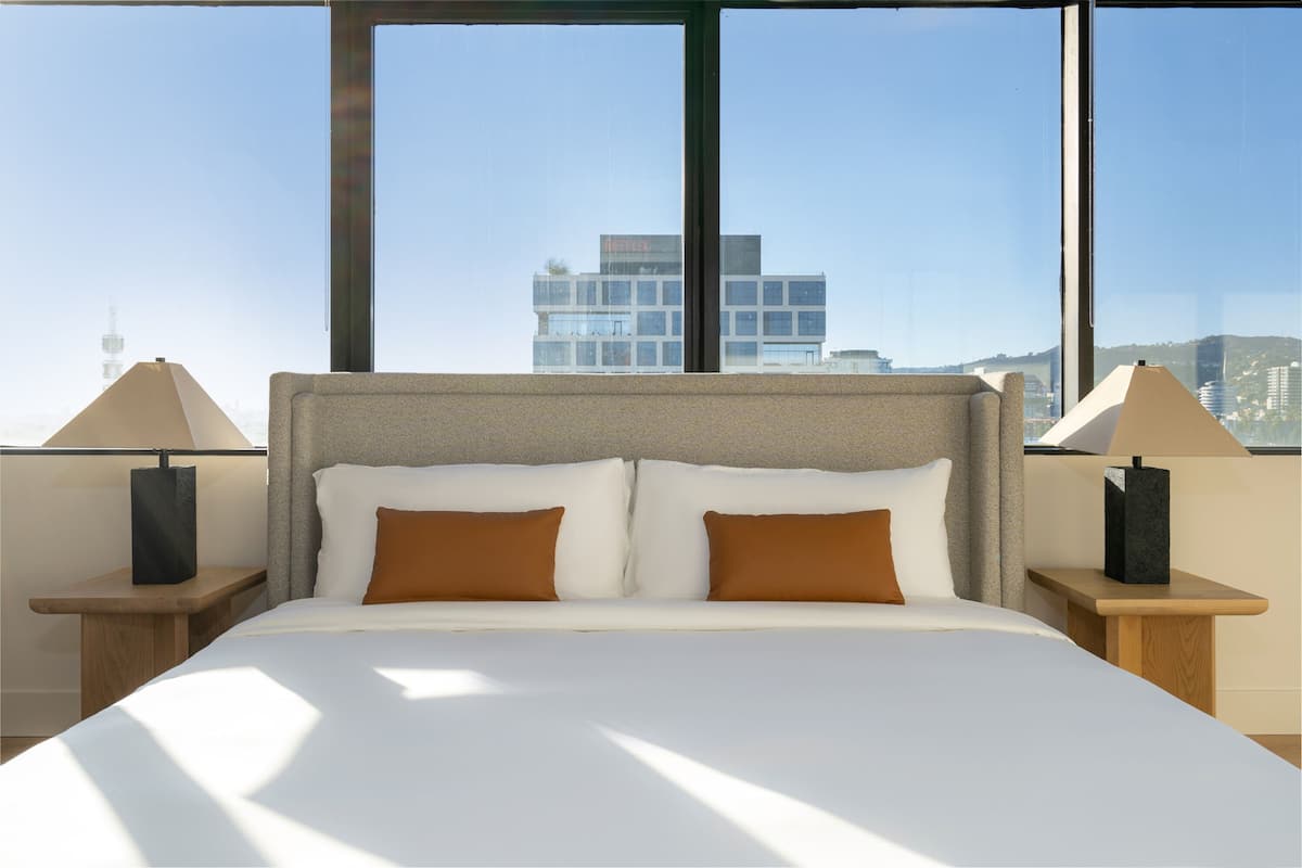 A hotel room with a large bed, two nightstands, and a view of a cityscape through large windows.