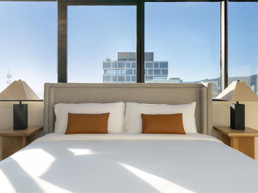 Habitación de hotel con una cama grande, dos mesitas de noche y vistas a un paisaje urbano a través de amplias ventanas.