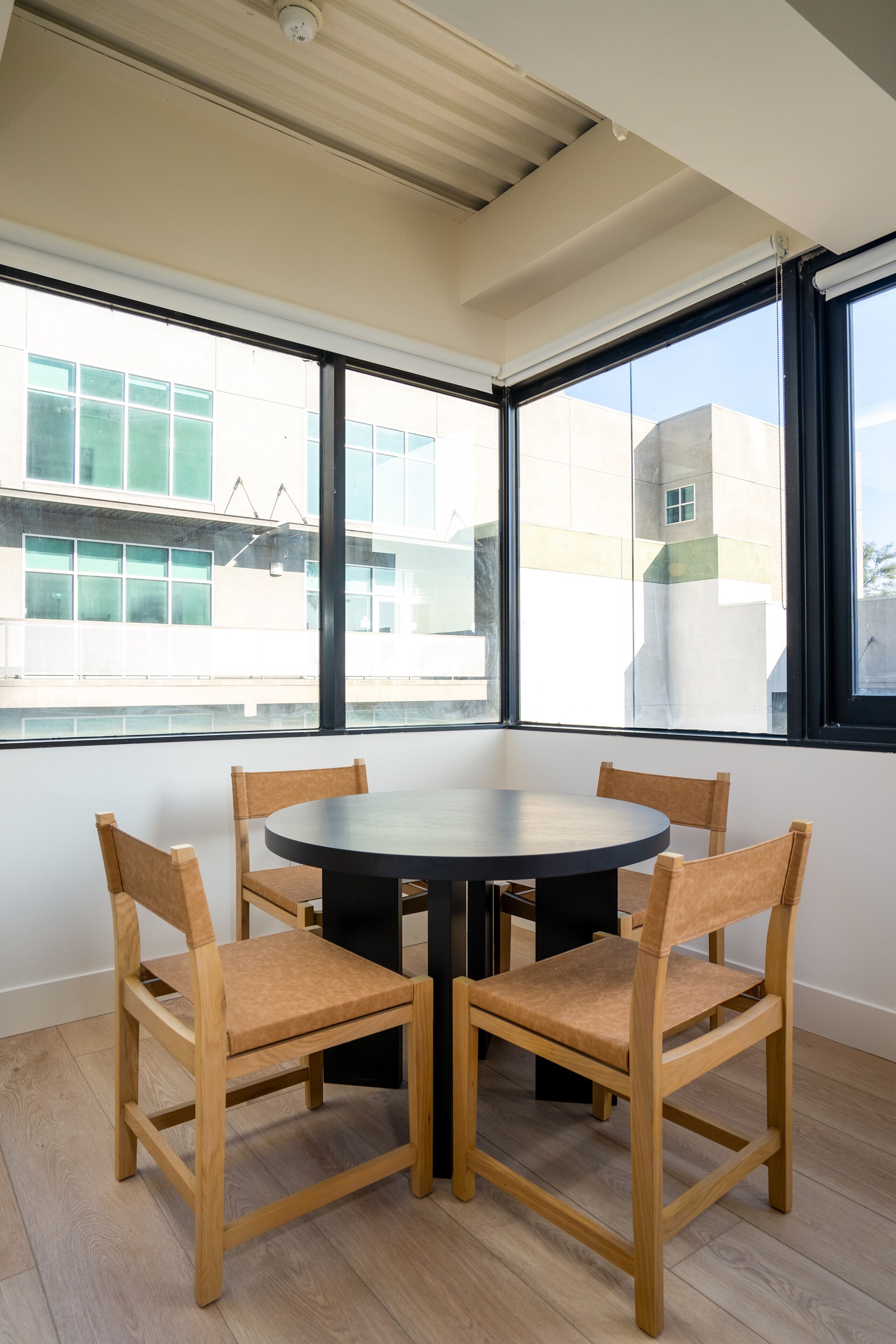 Una habitación con una mesa redonda y cuatro sillas de madera, ventanas amplias con vistas a edificios y un techo con una lámpara.
