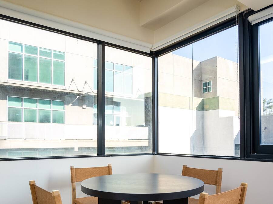A room with a round table and four wooden chairs, large windows with a view of buildings, and a ceiling with a light fixture.