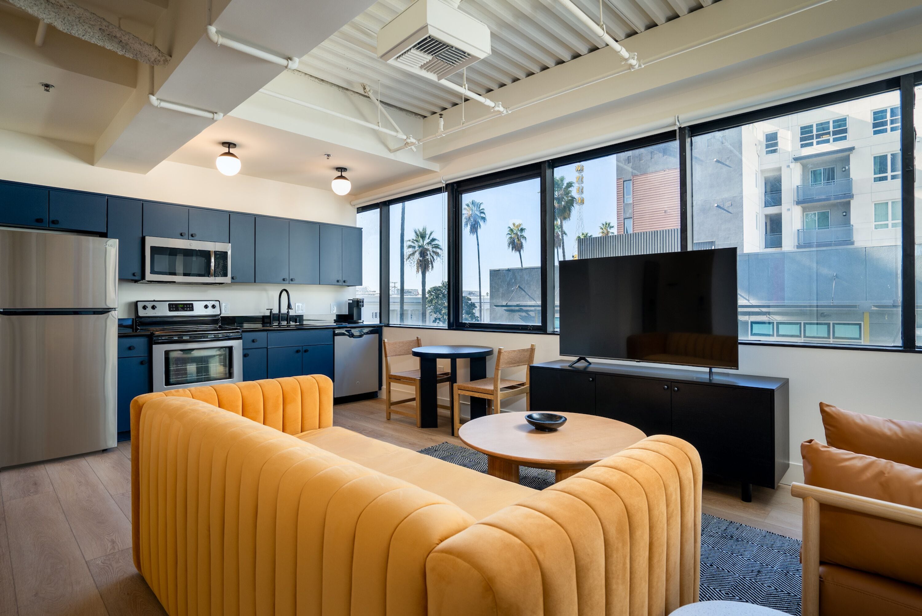 A modern living room with a sofa, a dining table, and a TV. There is a kitchen area with appliances and cabinets.