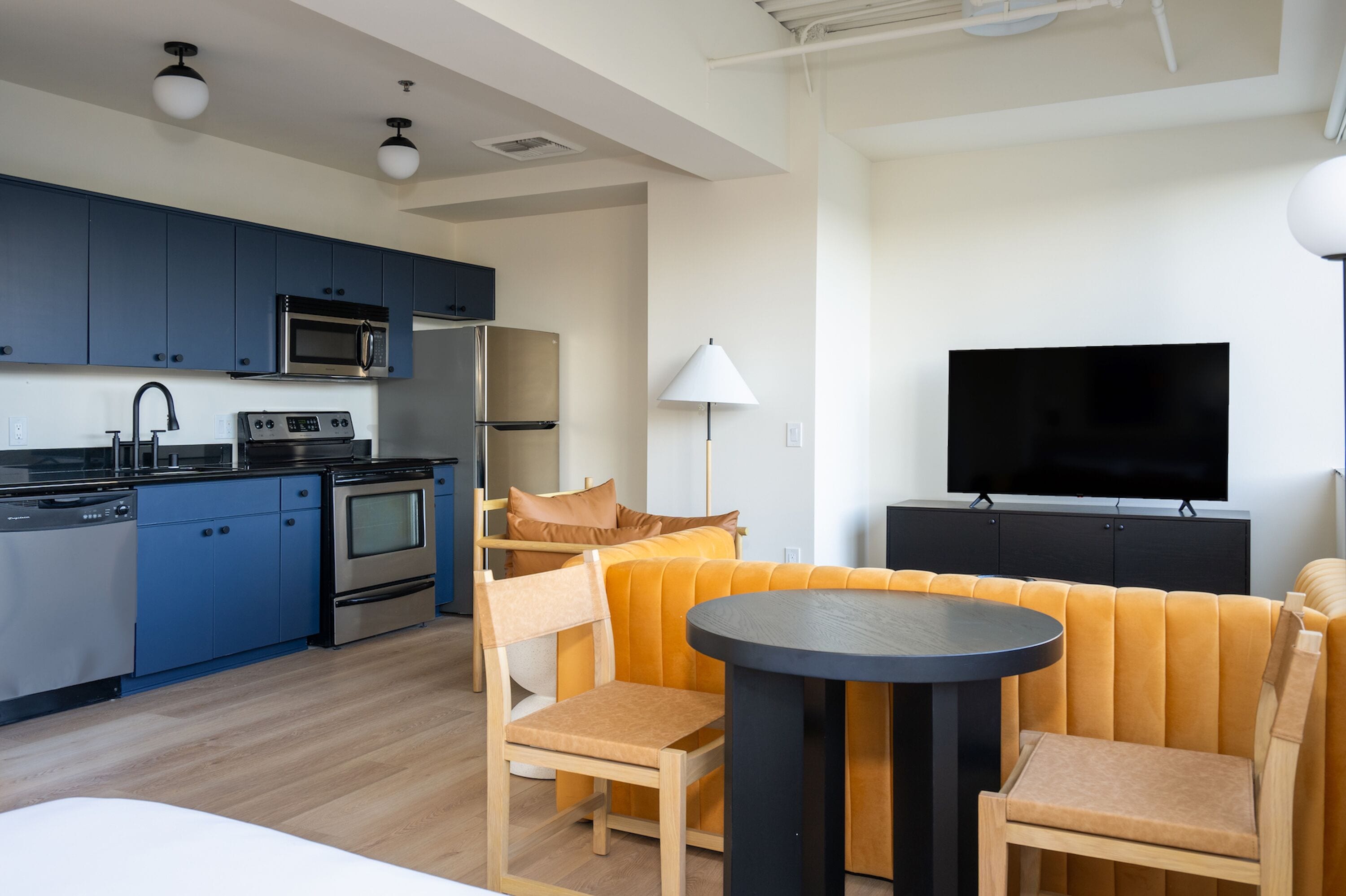 A modern kitchen with blue cabinets, a microwave, and a stove. There is a dining area with a round table and wooden chairs. A flat-screen TV is mounted on the wall.