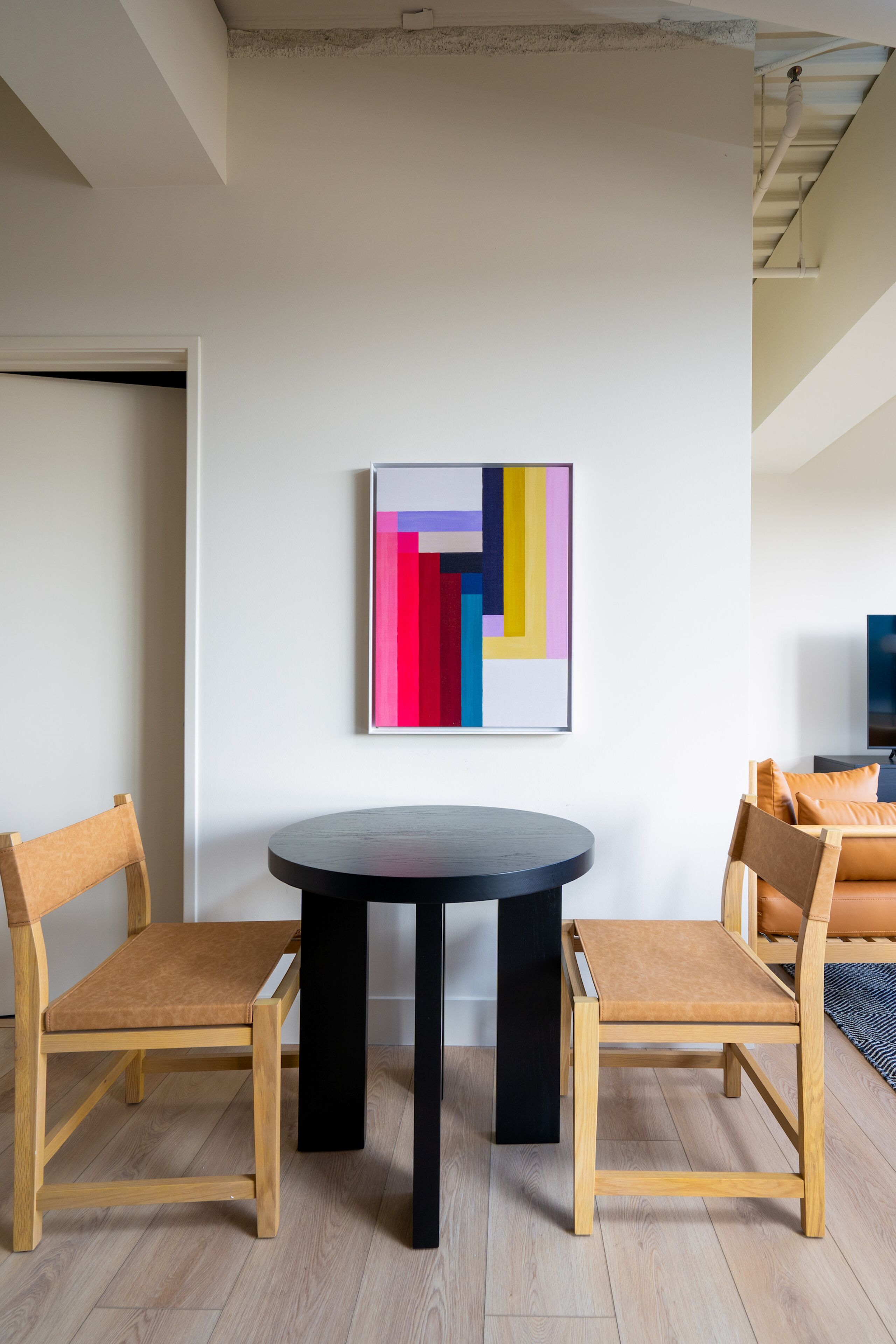 A modern living room with a round table, two wooden chairs, a colorful abstract painting, and a sofa with orange cushions.