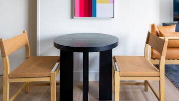 A modern living room with a round table, two wooden chairs, a colorful abstract painting, and a sofa with orange cushions.