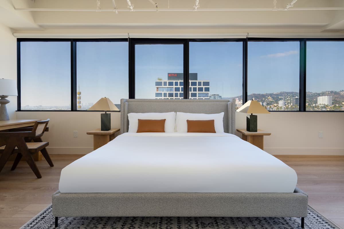 A modern hotel room with a large bed, two bedside lamps, a desk, and a chair. The room features large windows with a city view.