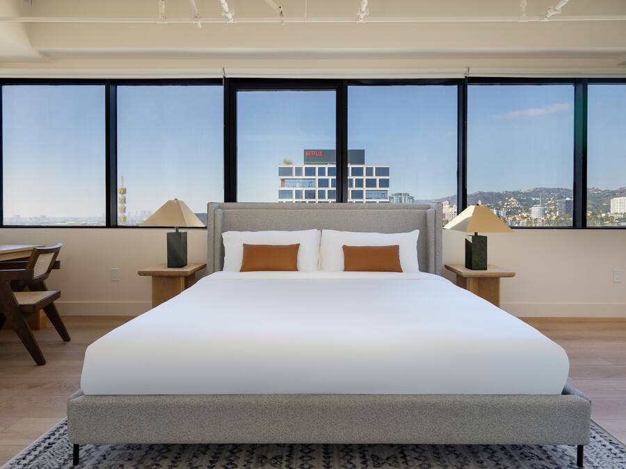 Una moderna camera d'albergo con un letto grande, due lampade ai comodini, una scrivania e una poltrona. La stanza ha ampie finestre con vista sulla città.