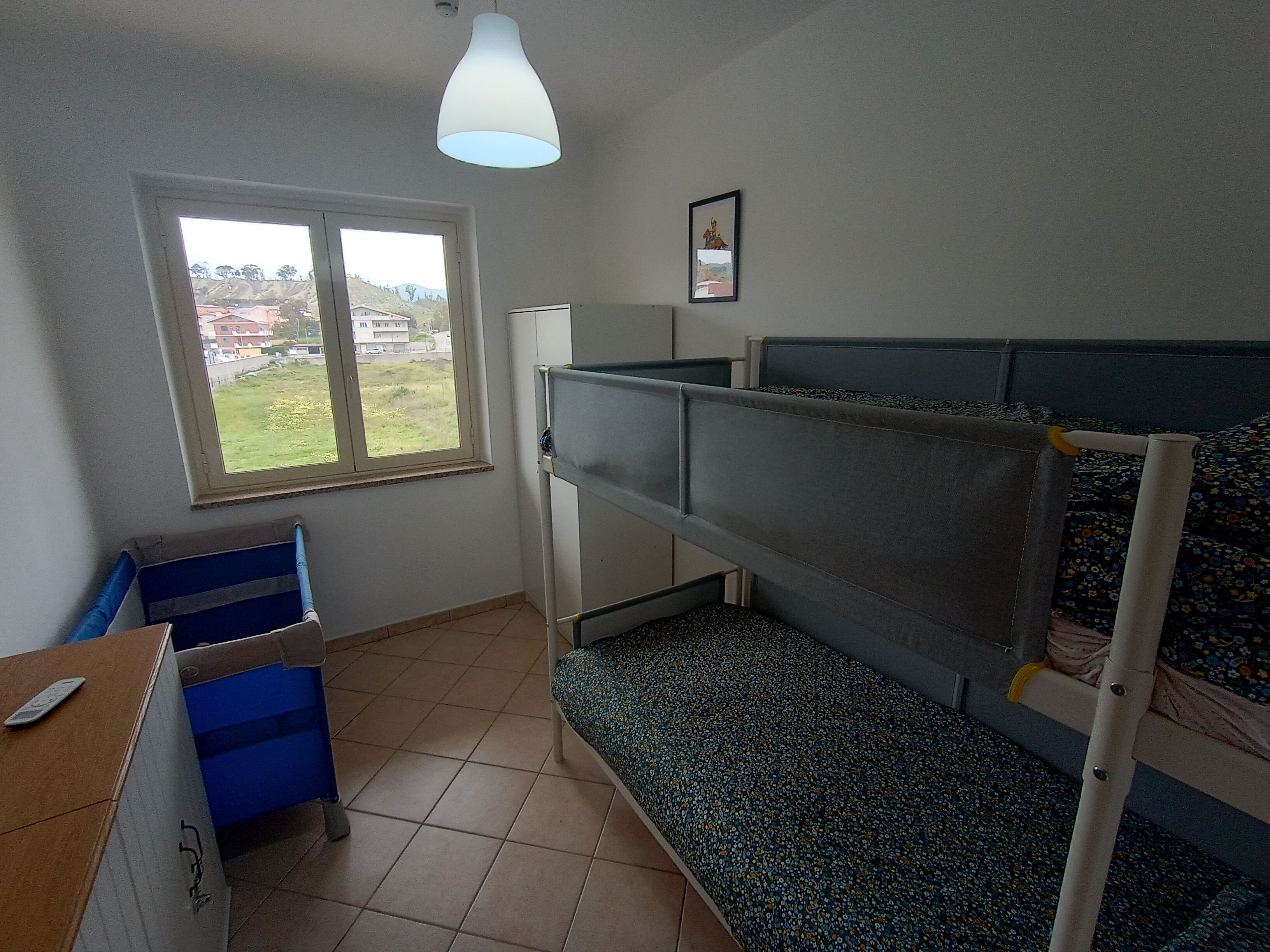 Een slaapzaal met stapelbedden, een bureau, een stoel, een raam met uitzicht op gebouwen en een plafondlamp.