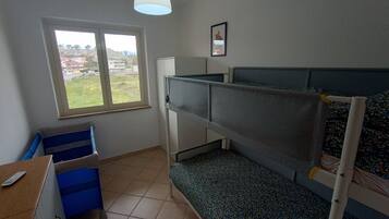 Een slaapzaal met stapelbedden, een bureau, een stoel, een raam met uitzicht op gebouwen en een plafondlamp.