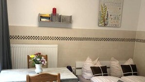 Ein kleiner, ordentlich eingerichteter Raum mit einem Bett, einem Tisch und einem Stuhl. Unter dem Tisch steht ein Heizkörper und an der Wand hängt ein Poster.